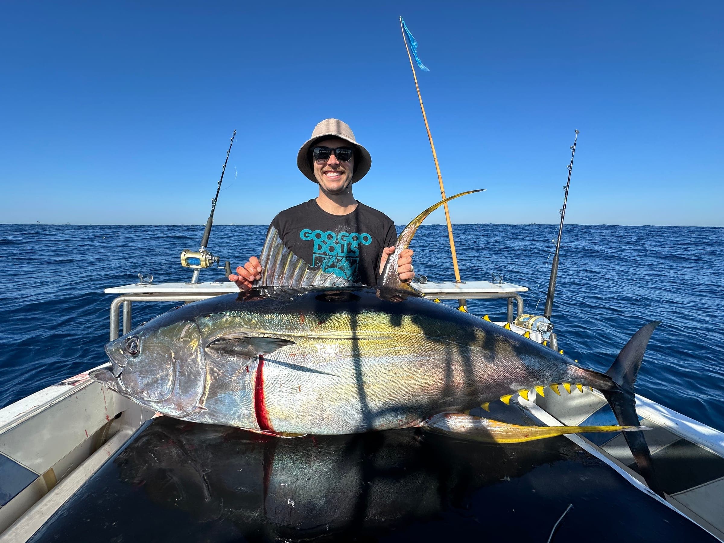 An angler standing behind a large yellowfin tuna laid across the gunwale under a clear sky.