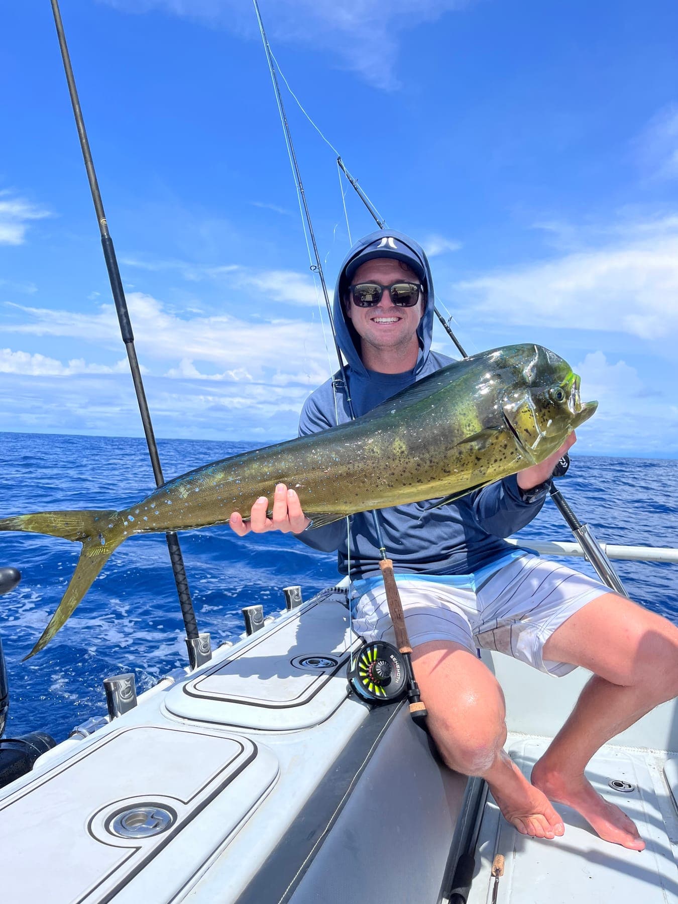 Garreth seated on the gunwale holding a dorado under a blue sky.