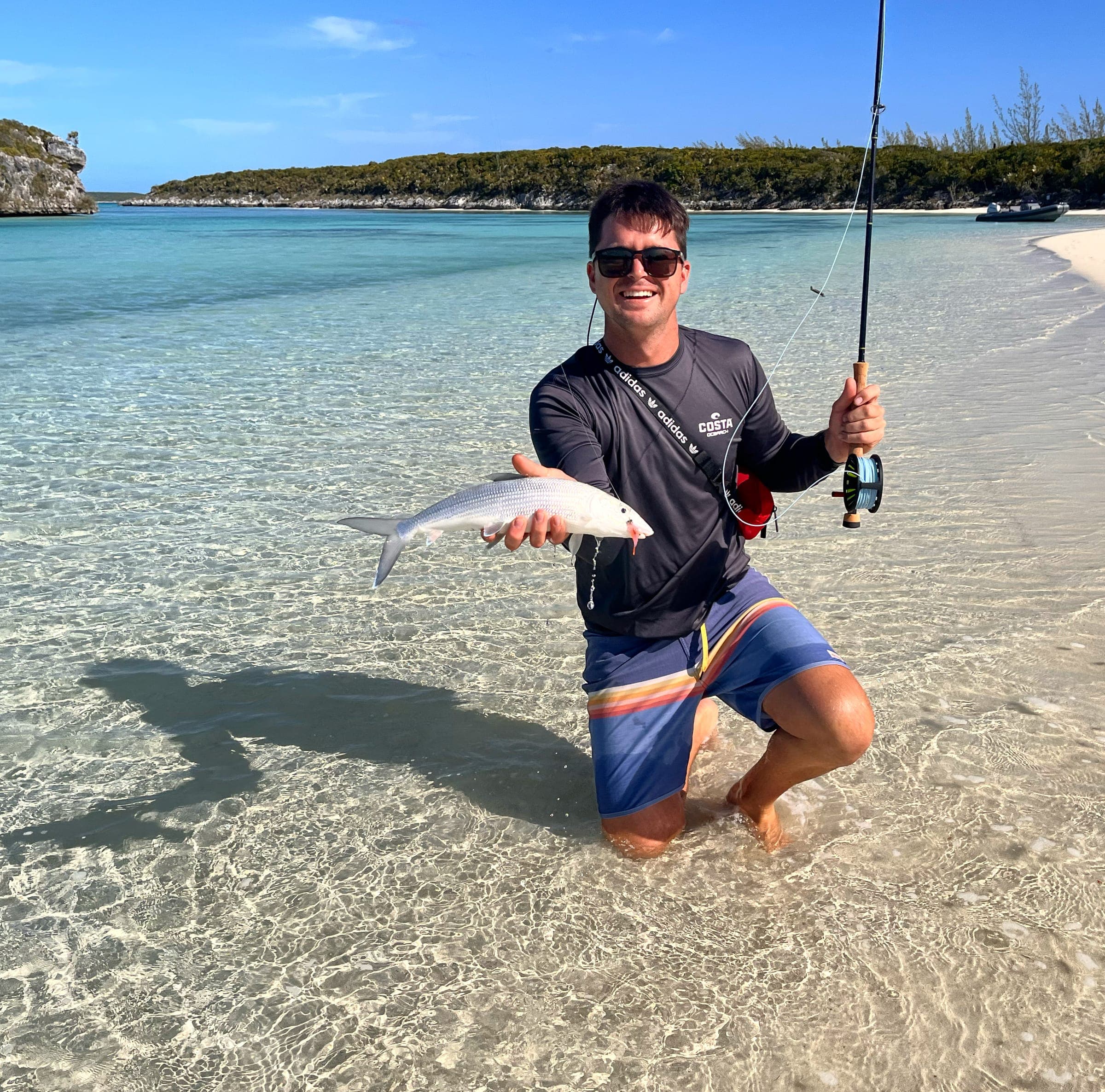 Garreth kneeling in clear shallow water off a white-sand beach, holding a silver fish.