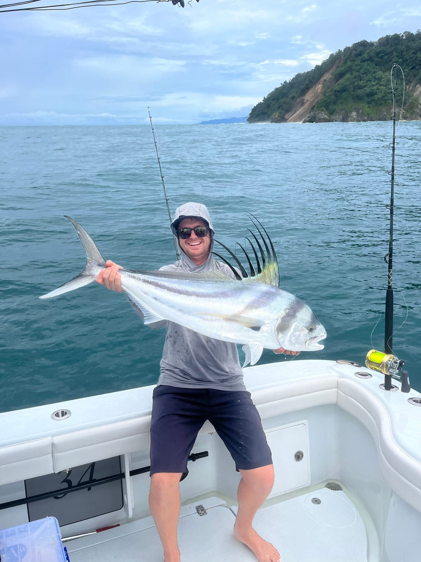 Garreth holding a roosterfish on deck, with a green headland behind.