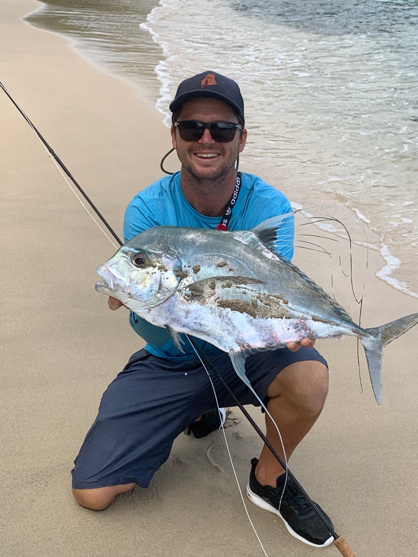 Garreth kneeling on a beach at the water’s edge holding a large trevally.