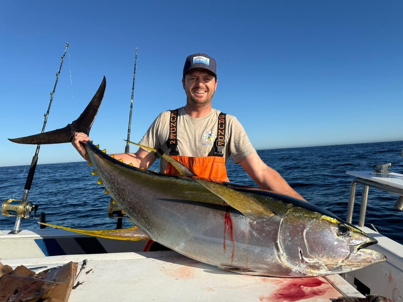 Garreth in orange bibs holding a large yellowfin tuna across the deck.