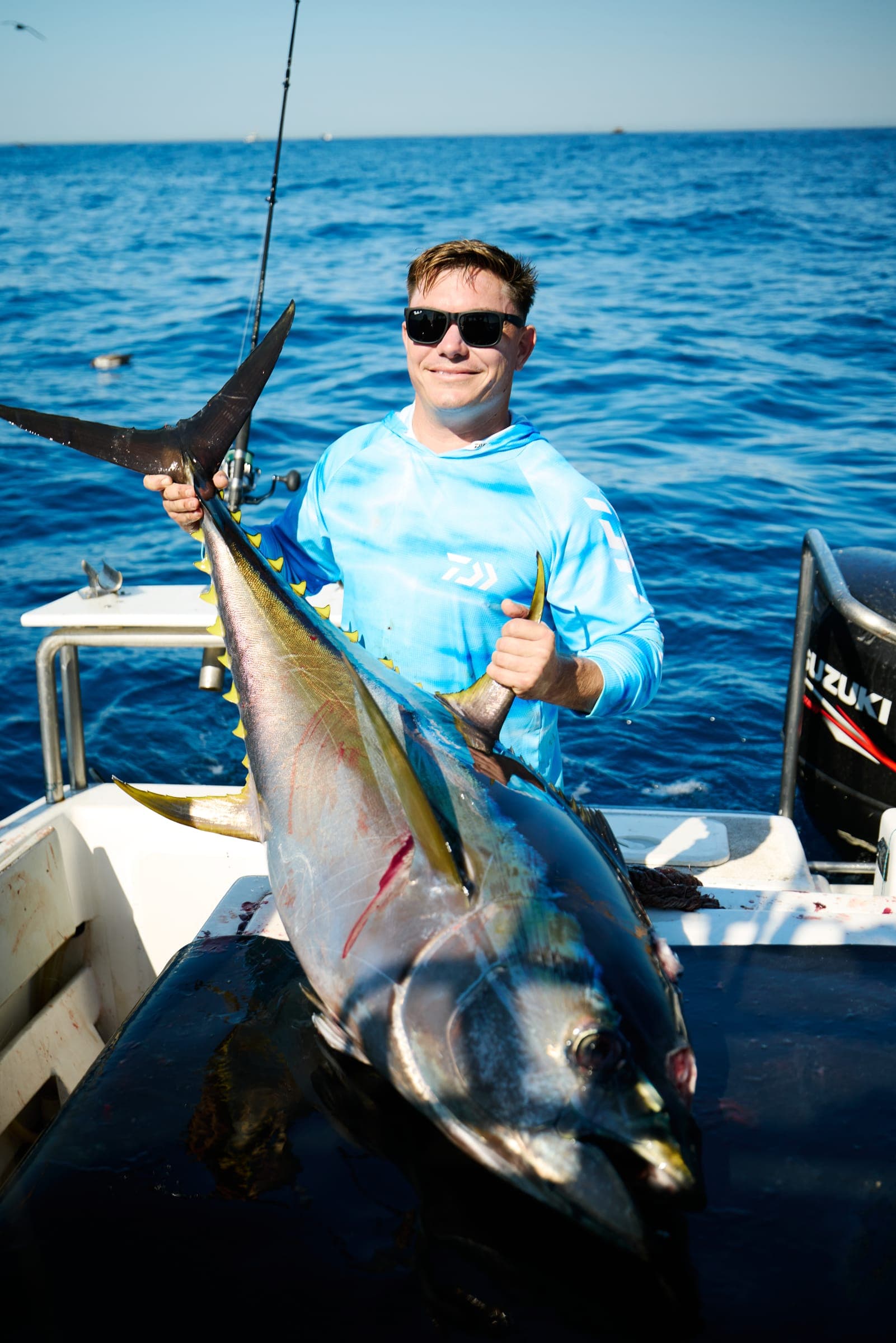 An angler in a blue shirt holding a tuna upright by the tail on the deck.