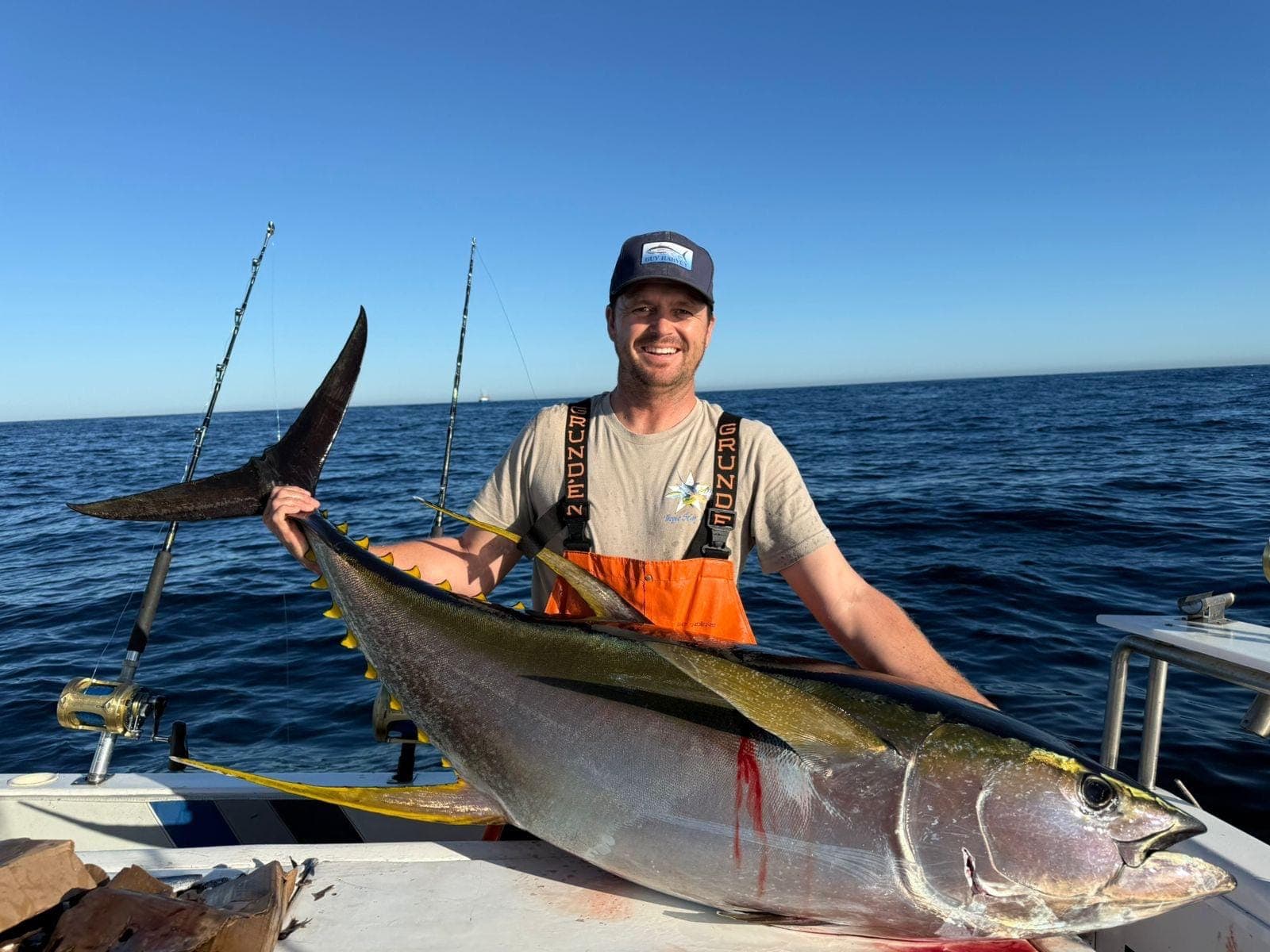 An angler in orange bibs smiling behind a yellowfin tuna laid across the rail.