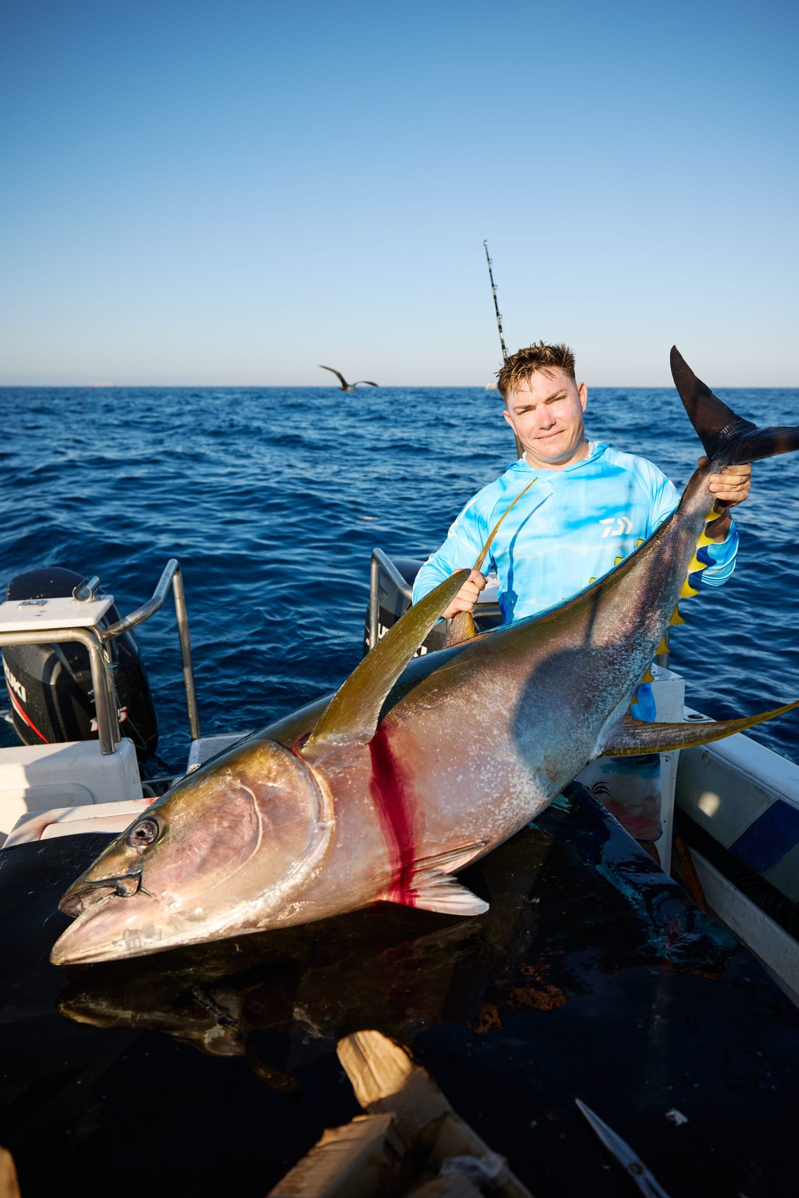 An angler kneeling on the deck holding a yellowfin tuna up by the tail, open blue water behind.