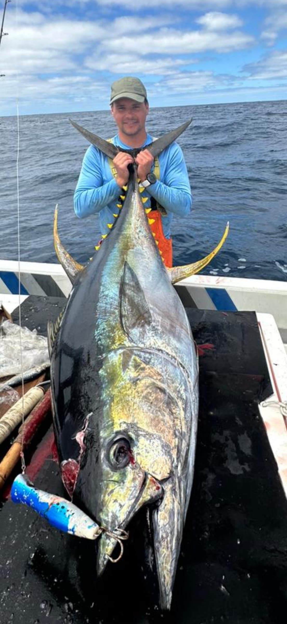 An angler holding a yellowfin tuna upright on the deck, with trolling lures beside it.