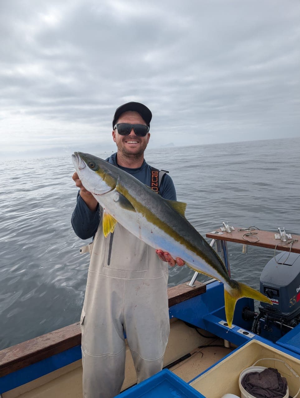 An angler holding a yellowtail on deck under a grey, overcast sky.