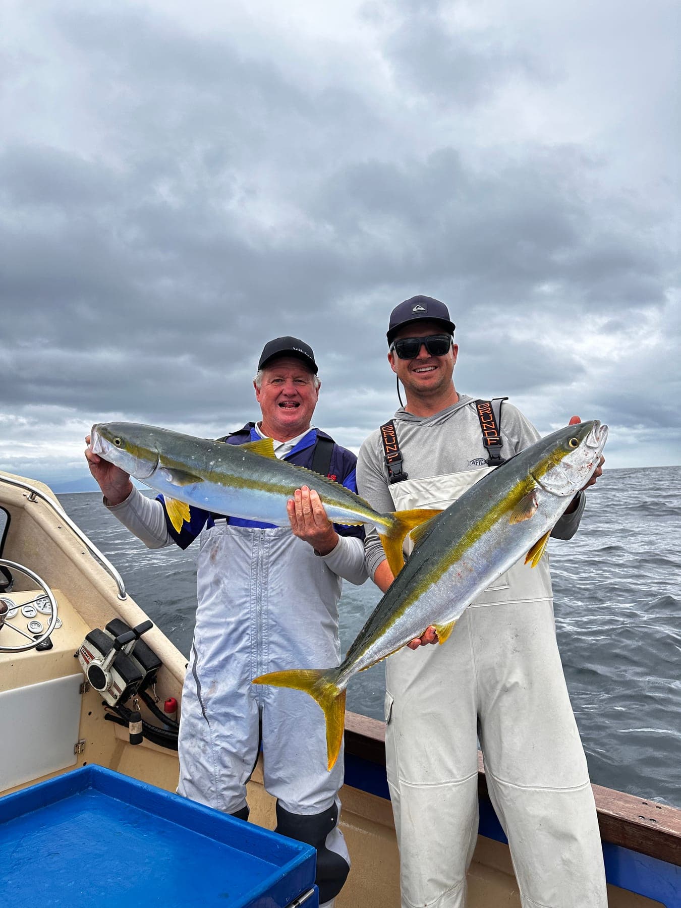 Two anglers in bibs each holding a yellowtail on an overcast day.