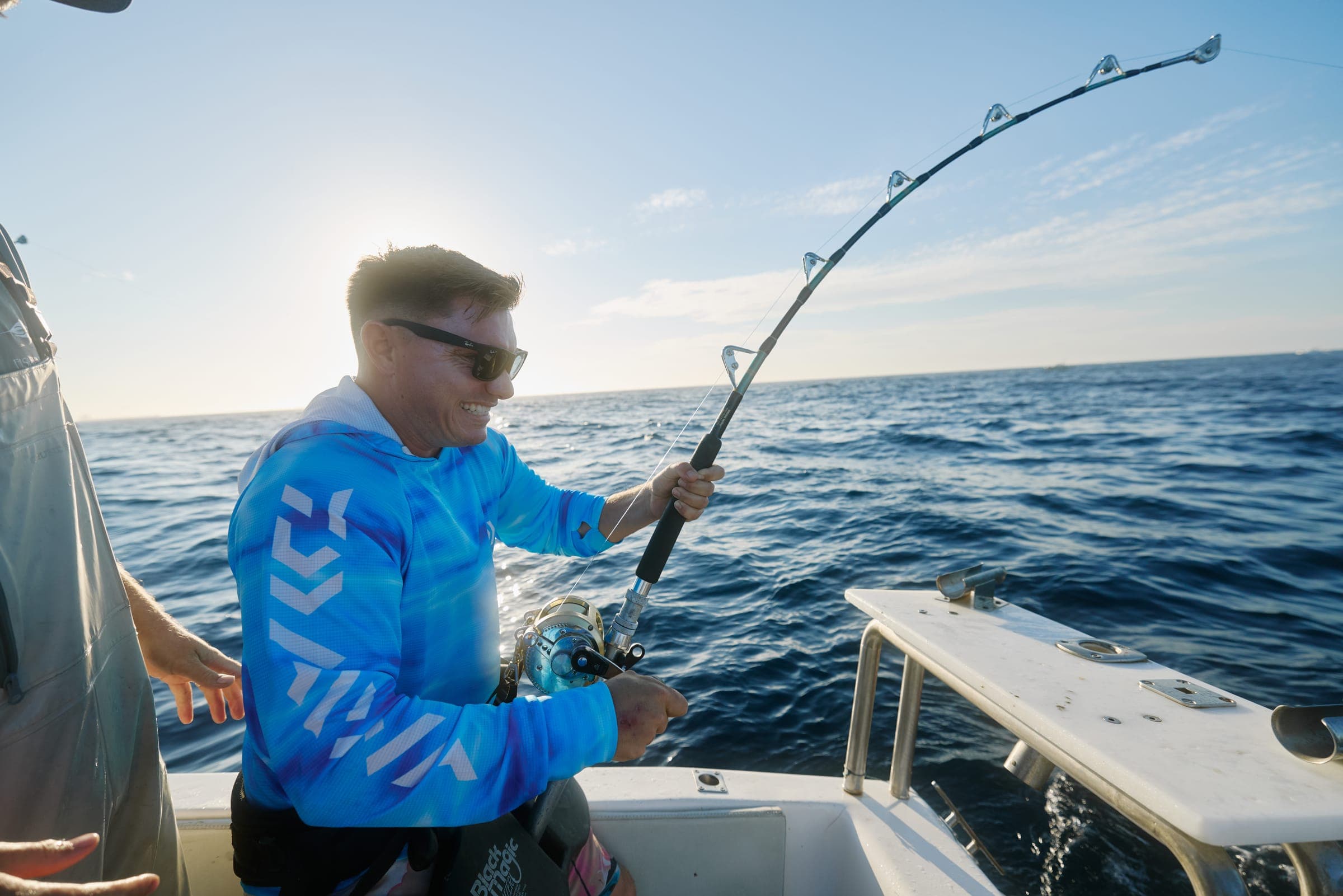 An angler braced against a deeply bent rod, backlit by low morning sun, fighting a fish alongside the boat.