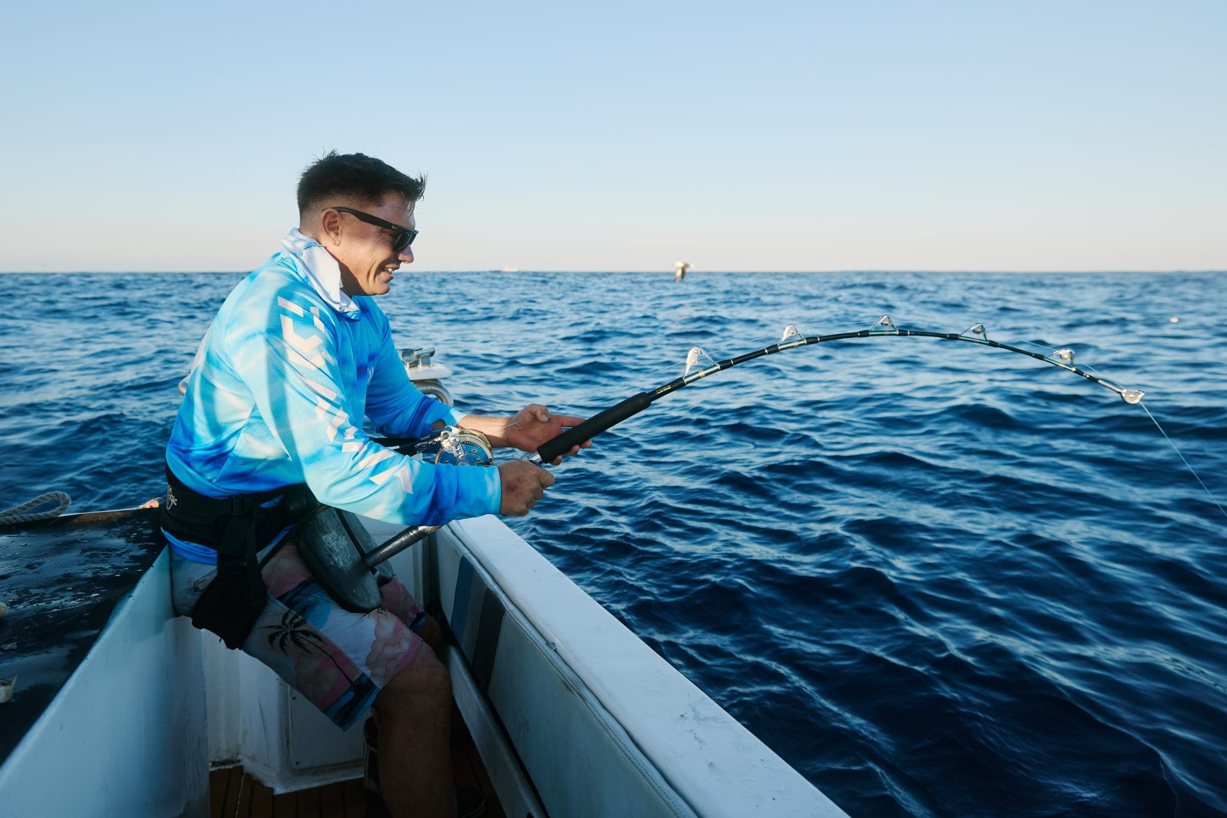 An angler seated on the gunwale, rod bent, working a fish on a calm sea.