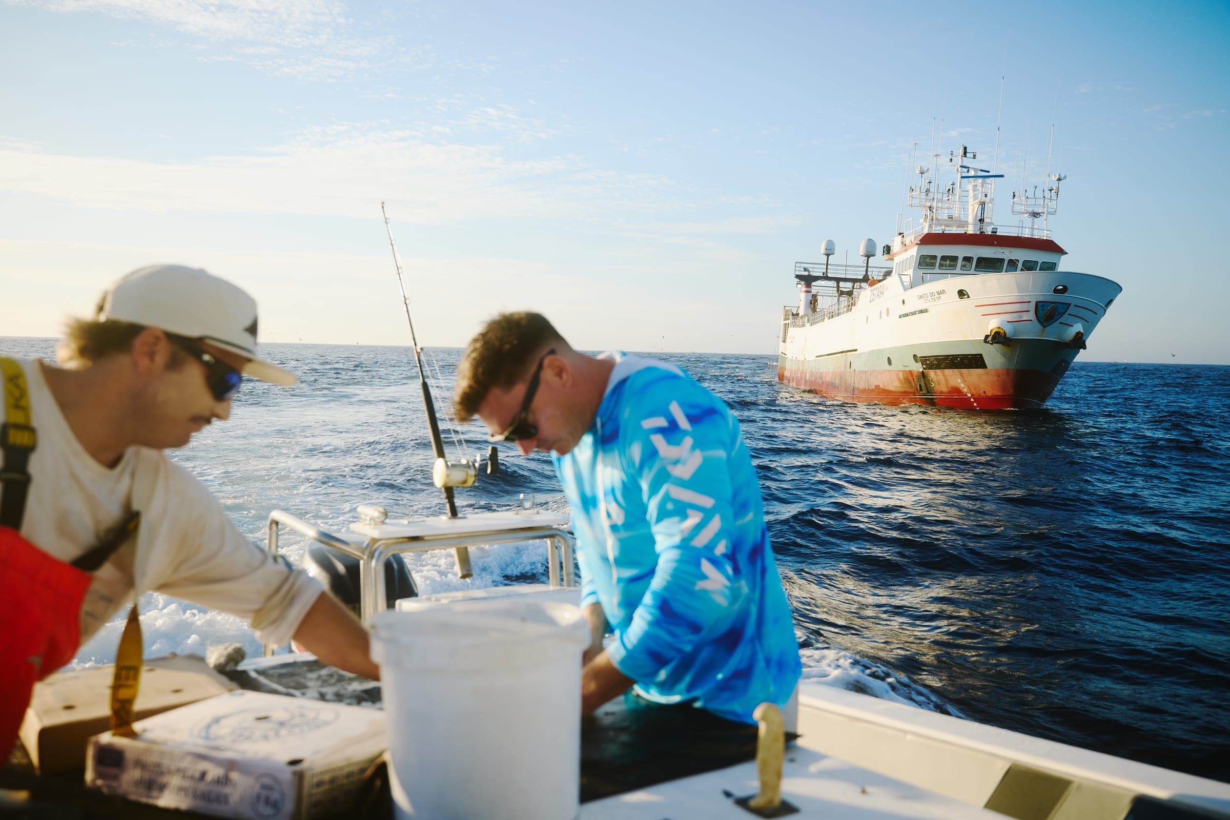Two crew working at the transom with a large fishing vessel lying off in the background.