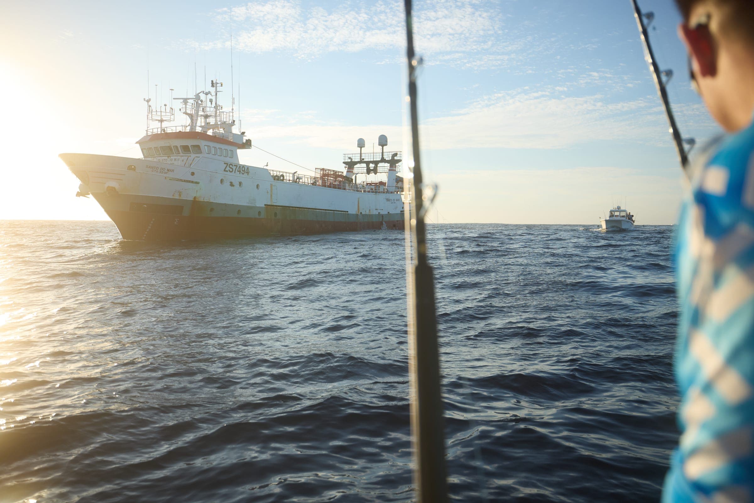 A trawler lying off in first light, framed between two rod tips from the deck of Benguela.