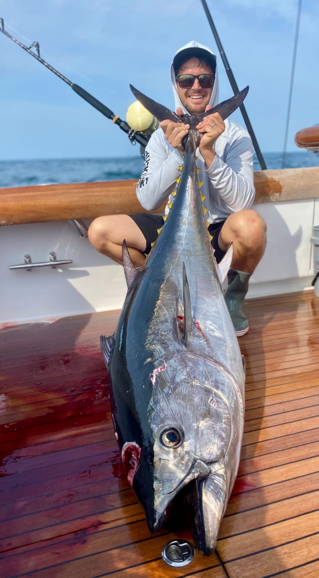 Captain Garreth crouched behind a very large tuna laid on the teak deck of a sportfishing boat, holding its tail upright.