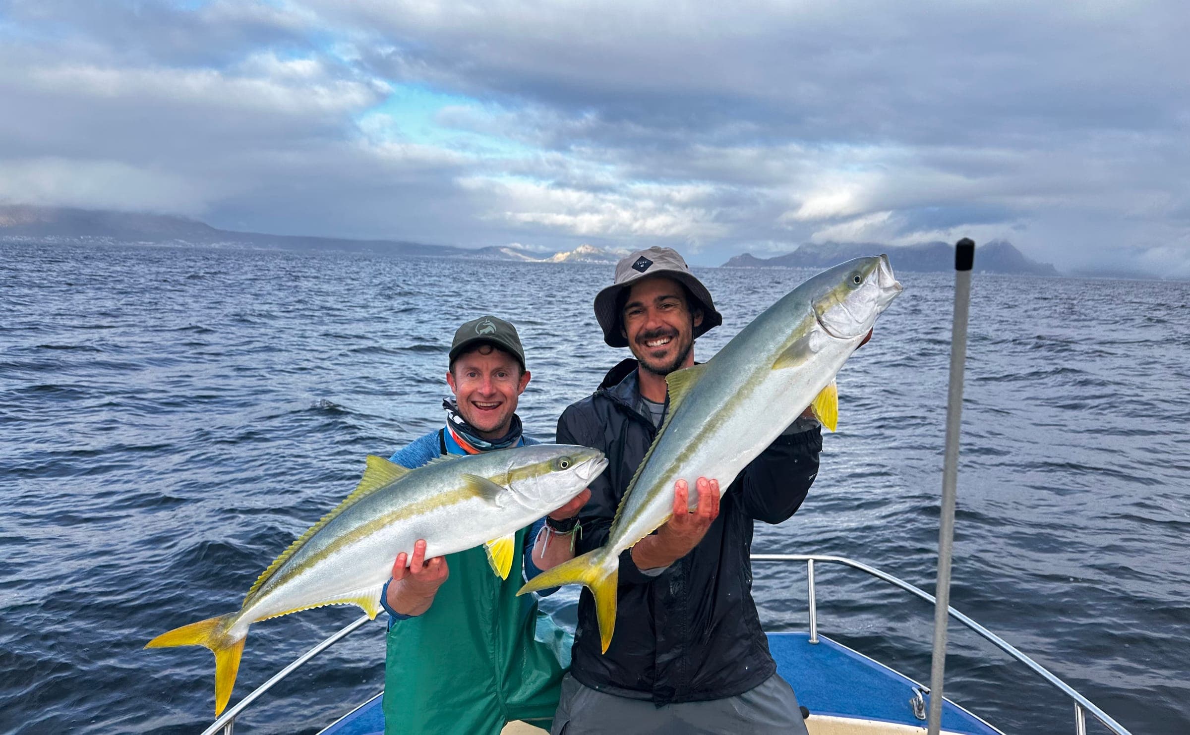 Two anglers each holding a yellowtail, with grey water and mountains behind.